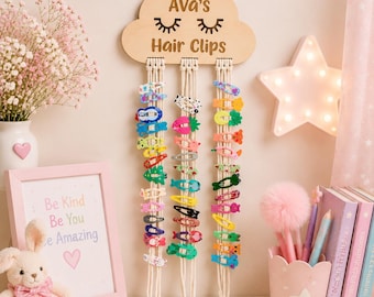 May include: A light-colored wooden cloud-shaped wall hanging with the engraved text Ava's Hair Clips and closed eye icons. Three long macrame cords hang from the cloud, each holding a vertical row of colorful, patterned metal hair clips.