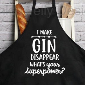 Puede incluir: Delantal negro con el texto blanco "I MAKE GIN DISAPPEAR WHAT'S your superpower?". Un pan y utensilios de madera son visibles en el fondo. El delantal tiene una correa para el cuello y lazos en la cintura.