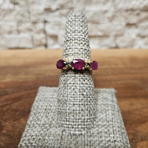 May include: A gold ring featuring three oval-cut, deep red gemstones, flanked by two smaller green stones. The ring is displayed on a gray ring holder against a neutral background. The gemstones are the focal point of this jewelry piece.