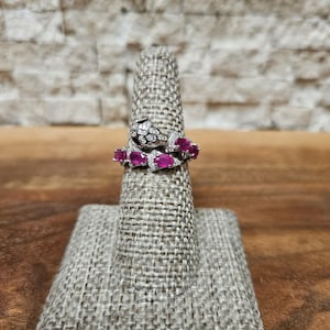 May include: Two rings displayed on a ring holder. One ring is a snake design with white stones and a black stone for the eye. The other ring has oval-shaped, deep pink stones and small white stones.