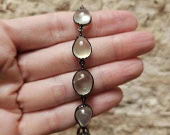 Puede incluir: Un brazalete con cinco cuentas de vidrio transparente en forma de lágrima engastadas en biseles de metal negro. El brazalete está en un cordón negro.