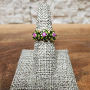 May include: A gold ring featuring three pink gemstones surrounded by green gemstones. The ring is displayed on a gray ring holder against a neutral background. The gemstones are set in a floral pattern.
