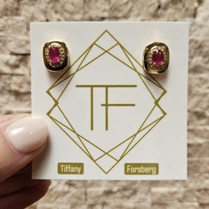 May include: A pair of gold stud earrings featuring oval-cut pink rubies surrounded by small clear stones. The earrings are mounted on a white display card with a gold geometric logo and the text TF, Tiffany, and Forsberg printed in gold.