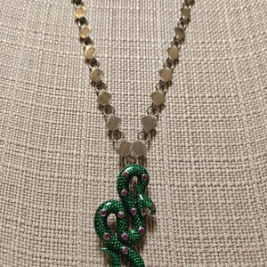 May include: A silver necklace featuring a green snake pendant with red detailing. The pendant hangs from a chain with silver and gold-coloured links. The necklace is presented against a textured, neutral-toned backdrop.
