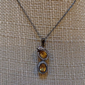 Op de afbeelding: Een zilveren ketting met een hanger met twee ovale stenen. De grotere steen is licht amberkleurig en de kleinere steen is donker amberkleurig. Beide stenen zijn omgeven door kleinere heldere stenen.