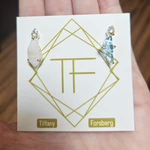 Peut inclure: Une paire de boucles d'oreilles dorées avec des pierres précieuses, présentées sur une carte blanche. Les boucles d'oreilles comportent une pierre précieuse transparente en haut et une pierre précieuse facettée plus grande de couleur claire. La carte porte le texte "Tiffany Forsberg".