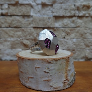 May include: A silver ring with a geometric faceted design set with pink gemstones. The ring is displayed on a wooden surface.