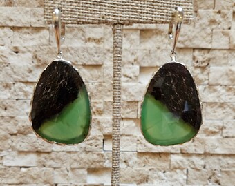 May include: A pair of dangle earrings with a silver-colored metal setting. Each earring features a large, teardrop-shaped stone, with a dark brown top half and a light green bottom half. The earrings are displayed on a neutral-colored stand.
