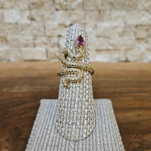 May include: A gold ring shaped like a coiled snake wrapping around the finger. The snake body is covered in small, clear pavé-set stones, and the head features a single, pear-shaped, deep pink gemstone set as the eye of the serpent.
