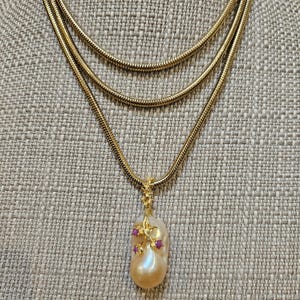 May include: A gold-toned necklace with three strands and a pendant. The pendant features a large, pear-shaped pearl with small red gemstones and gold accents. The necklace is displayed on a textured, neutral-colored surface.