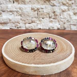 May include: A pair of gemstone earrings. Each earring features a large, oval-shaped, clear quartz stone with golden inclusions, surrounded by a halo of small red and blue gemstones. The earrings are set on a wooden surface.
