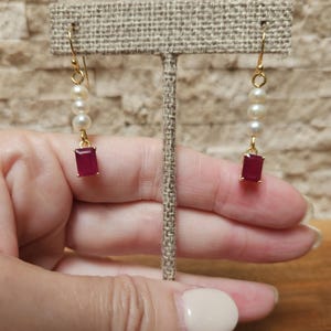 May include: A pair of gold-toned earrings with a drop design. Each earring features a rectangular, deep red gemstone at the bottom, with three small pearls above it. The earrings are displayed on a neutral-colored jewelry stand.