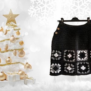 May include: A black crochet skirt with a white and gray geometric pattern. The skirt is hanging on a black hanger against a white background with a white Christmas tree decorated with gold ornaments.