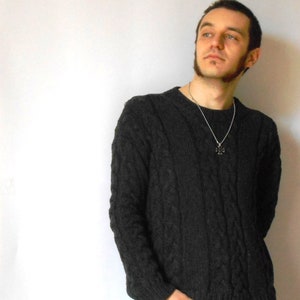 May include: A man wearing a dark gray cable knit sweater with a silver chain necklace and a cross pendant.
