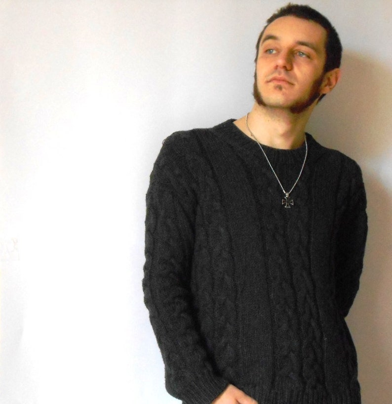 May include: A man wearing a dark gray cable knit sweater with a silver chain necklace and a cross pendant.