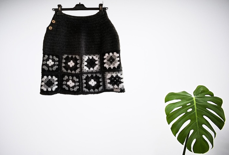 May include: A gray crochet skirt with a black and white geometric pattern. The skirt is hanging on a hanger against a white wall.