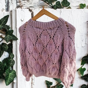 K&ouml;nnte beinhalten: Ein blassrosa gestrickter Pullover mit einem Rautenmuster und durchbrochenem Design. Der Pullover h&auml;ngt an einem Holzb&uuml;gel vor einem wei&szlig;en Hintergrund.