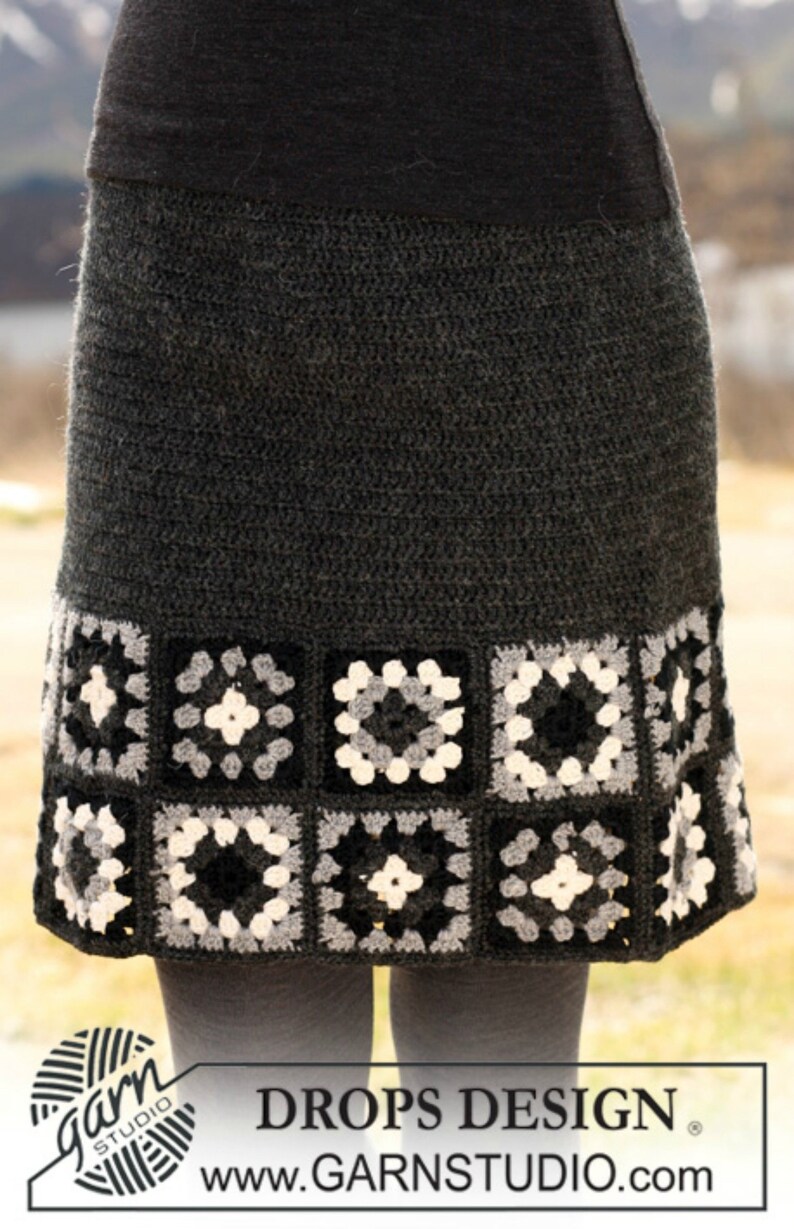May include: A gray crochet skirt with a black, white, and gray granny square border. The skirt is made with a simple stitch and has a fitted waist. The skirt is shown on a model wearing black tights.