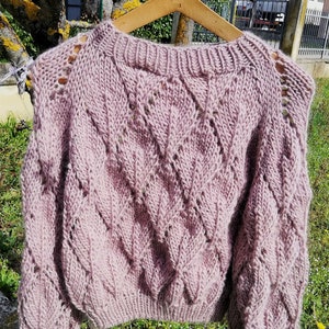 K&ouml;nnte beinhalten: Ein weicher, rosa gestrickter Pullover mit einem Zopfmuster und durchbrochenen Details. Der Pullover hat eine lockere Passform und lange &Auml;rmel.