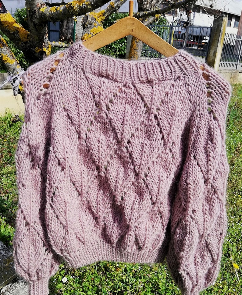 K&ouml;nnte beinhalten: Ein weicher, rosa gestrickter Pullover mit einem Zopfmuster und durchbrochenen Details. Der Pullover hat eine lockere Passform und lange &Auml;rmel.