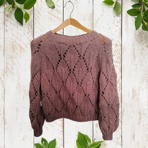 K&ouml;nnte beinhalten: Ein mauvefarbener Strickpullover mit einem durchbrochenen Rautenmuster. Der Pullover h&auml;ngt an einem Holzb&uuml;gel vor einem wei&szlig;en Holz Hintergrund.