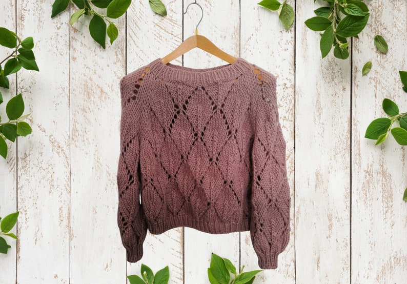 K&ouml;nnte beinhalten: Ein mauvefarbener Strickpullover mit einem durchbrochenen Rautenmuster. Der Pullover h&auml;ngt an einem Holzb&uuml;gel vor einem wei&szlig;en Holz Hintergrund.