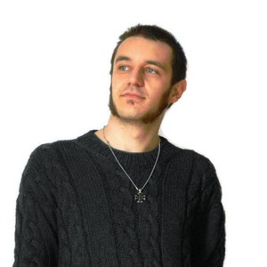 May include: A man wearing a dark gray cable knit sweater with a silver chain necklace and a small cross pendant.