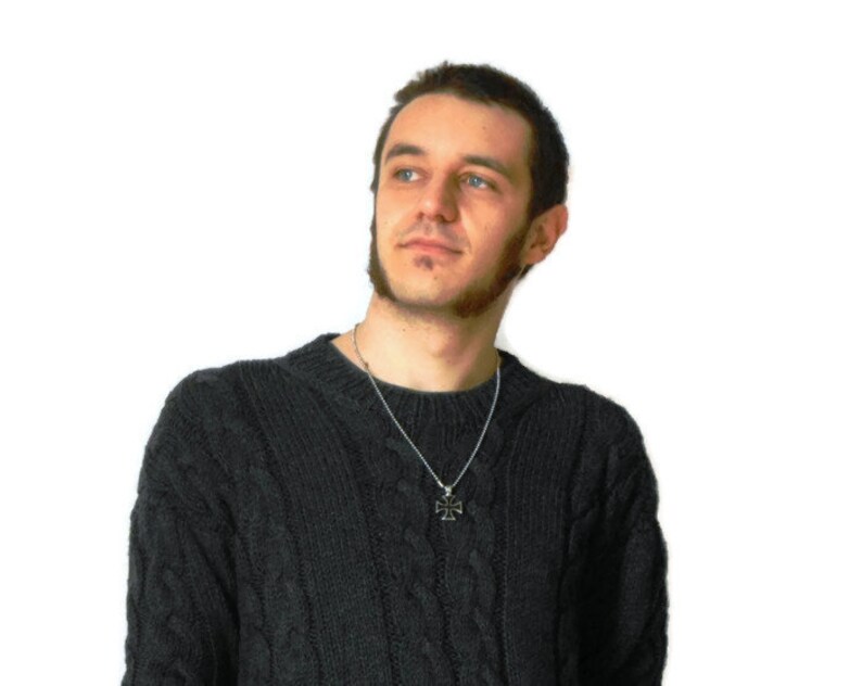 May include: A man wearing a dark gray cable knit sweater with a silver chain necklace and a small cross pendant.