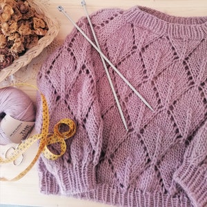 K&ouml;nnte beinhalten: Nahaufnahme eines rosa gestrickten Pullovers mit Rautenmuster. Zwei Stricknadeln liegen auf dem Pullover. Ein Ma&szlig;band ist ebenfalls auf dem Bild zu sehen.