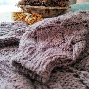 K&ouml;nnte beinhalten: Nahaufnahme eines weichen, mauvefarbenen Strickpullovers mit einem Zopfmuster. Ein gelbes Ma&szlig;band und Stricknadeln sind im Hintergrund zu sehen.