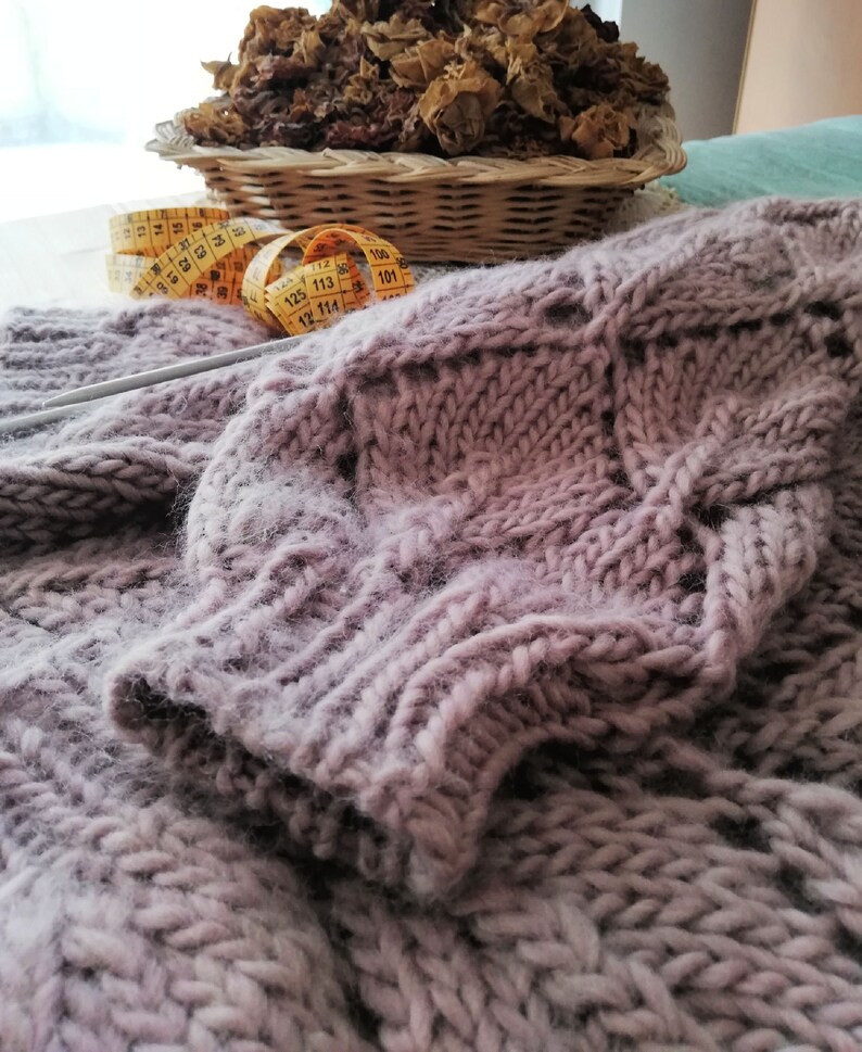 K&ouml;nnte beinhalten: Nahaufnahme eines weichen, mauvefarbenen Strickpullovers mit einem Zopfmuster. Ein gelbes Ma&szlig;band und Stricknadeln sind im Hintergrund zu sehen.