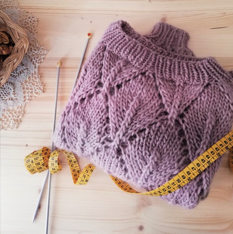 K&ouml;nnte beinhalten: Nahaufnahme eines lilafarbenen Strickpullovers mit einem Zopfmuster. Ein gelbes Ma&szlig;band ist um den Pullover gewickelt, und zwei Stricknadeln liegen auf einer Holzoberfl&auml;che.