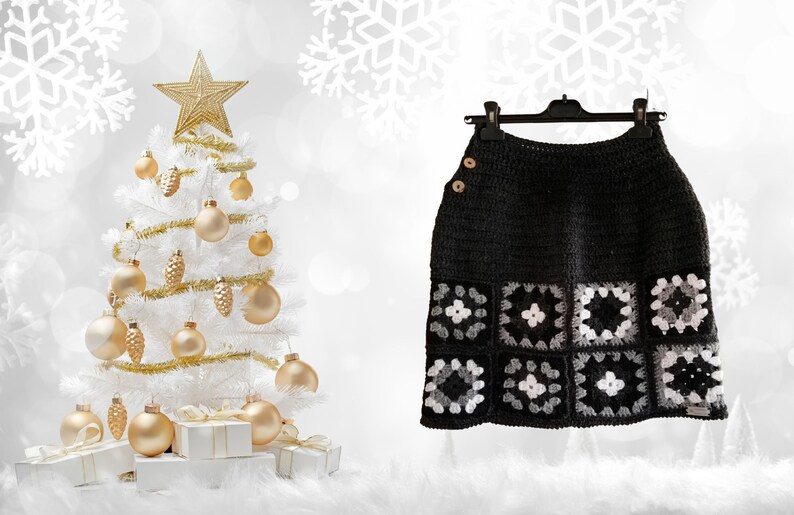 May include: A black crochet skirt with a white and gray geometric pattern. The skirt is hanging on a black hanger against a white background with a white Christmas tree decorated with gold ornaments.