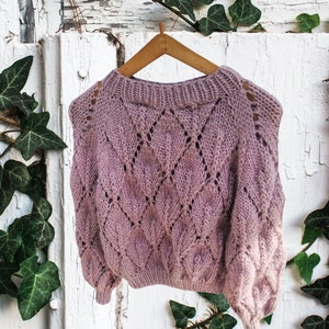K&ouml;nnte beinhalten: Ein blassrosa gestrickter Pullover mit einem Rautenmuster und durchbrochenem Design. Der Pullover h&auml;ngt an einem Holzb&uuml;gel vor einem wei&szlig;en Hintergrund.