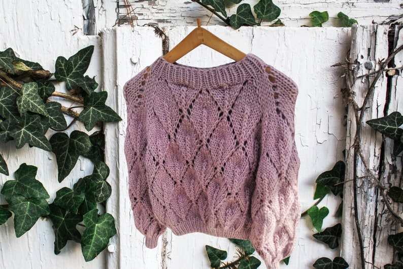K&ouml;nnte beinhalten: Ein blassrosa gestrickter Pullover mit einem Rautenmuster und durchbrochenem Design. Der Pullover h&auml;ngt an einem Holzb&uuml;gel vor einem wei&szlig;en Hintergrund.