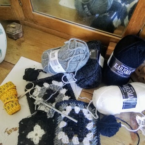 May include: Close-up of a crochet project with black, white, and gray yarn. The project is made up of granny squares. There are also balls of yarn in black, white, and gray, as well as a measuring tape and a crochet hook.