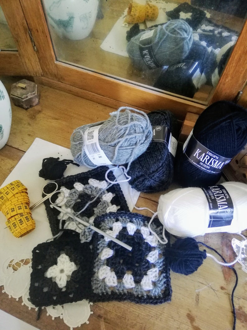 May include: Close-up of a crochet project with black, white, and gray yarn. The project is made up of granny squares. There are also balls of yarn in black, white, and gray, as well as a measuring tape and a crochet hook.