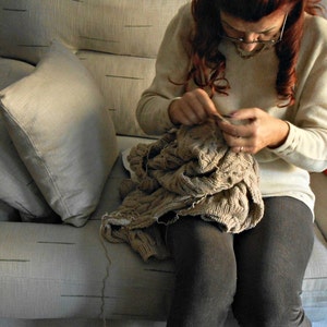 May include: A woman sits on a couch and knits a brown sweater. She is wearing a white sweater and has her hair pulled back.