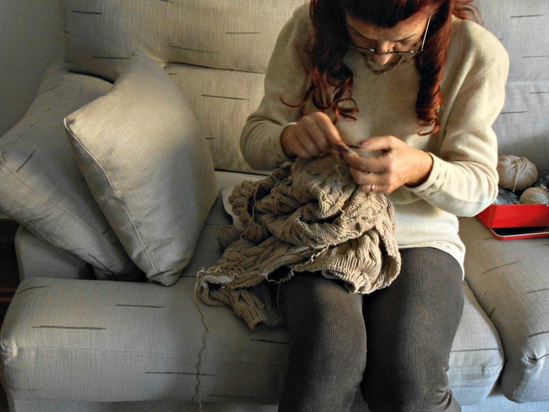 May include: A woman sits on a couch and knits a brown sweater. She is wearing a white sweater and has her hair pulled back.