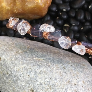 May include: A copper-colored hair barrette adorned with clear and purple gemstones. The barrette is wrapped with copper wire, and the gemstones are irregularly shaped. The barrette is displayed on a bed of black beans.