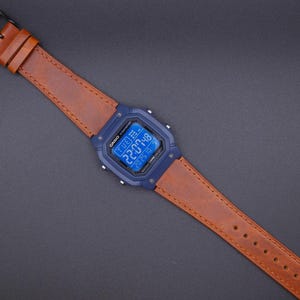 May include: A digital watch with a blue face and a brown leather strap. The watch displays the time and date. The strap has a black buckle and is stitched along the edges. The watch is a square shape.