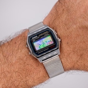 Könnte beinhalten: Eine silberne digitale Casio-Uhr mit Mesh-Armband zeigt die Uhrzeit 2:27:05 an. Das Zifferblatt hat einen blauen Rand und trägt die Aufschrift "CASIO ALARM CHRONO" und "WATER RESIST". Die Uhr befindet sich an einem Handgelenk.