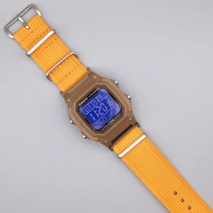 May include: A digital watch with a brown case and a bright yellow nylon strap. The watch face displays the time and date in blue. The strap is secured with a silver buckle.