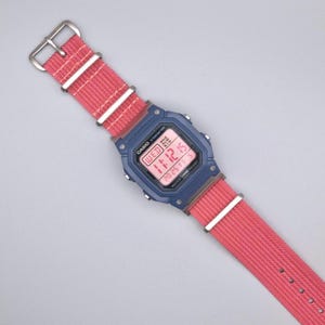 May include: A digital watch with a navy blue case and a pink nylon strap. The watch face displays the time, date, and day of the week in a digital format. The strap has white stripes and a silver buckle.