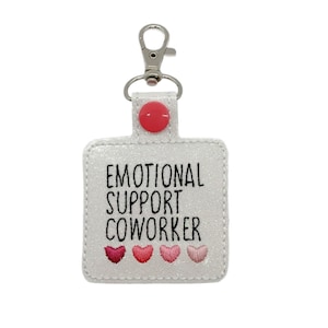 May include: A white glittery keychain with the text "EMOTIONAL SUPPORT COWORKER" in black. The keychain has a red button and four embroidered hearts in shades of pink. A silver-colored clasp is attached.