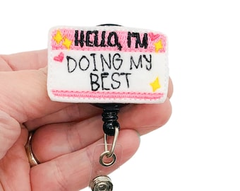 Puede incluir: Una insignia bordada blanca y rosa con el texto "Hello I'm Doing My Best" y estrellas amarillas. La insignia está unida a un carrete retráctil de insignia negro.