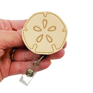 May include: A beige sand dollar shaped badge holder with a gold embroidered design. The badge holder is attached to a clear plastic retractable badge reel.