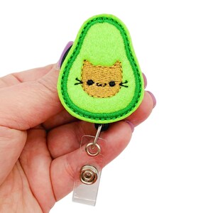 May include: A green avocado-shaped badge holder with a cute cat face embroidered on it. The badge holder has a clear plastic retractable cord.