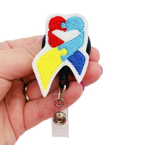May include: A white, blue, yellow, and red ribbon with a puzzle piece design. The ribbon is attached to a black retractable badge reel with a clear plastic badge holder.