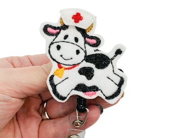 May include: White felt cow with black spots wearing a red and white nurse's hat with a red cross. The cow is holding a retractable badge reel with a clear plastic clip.